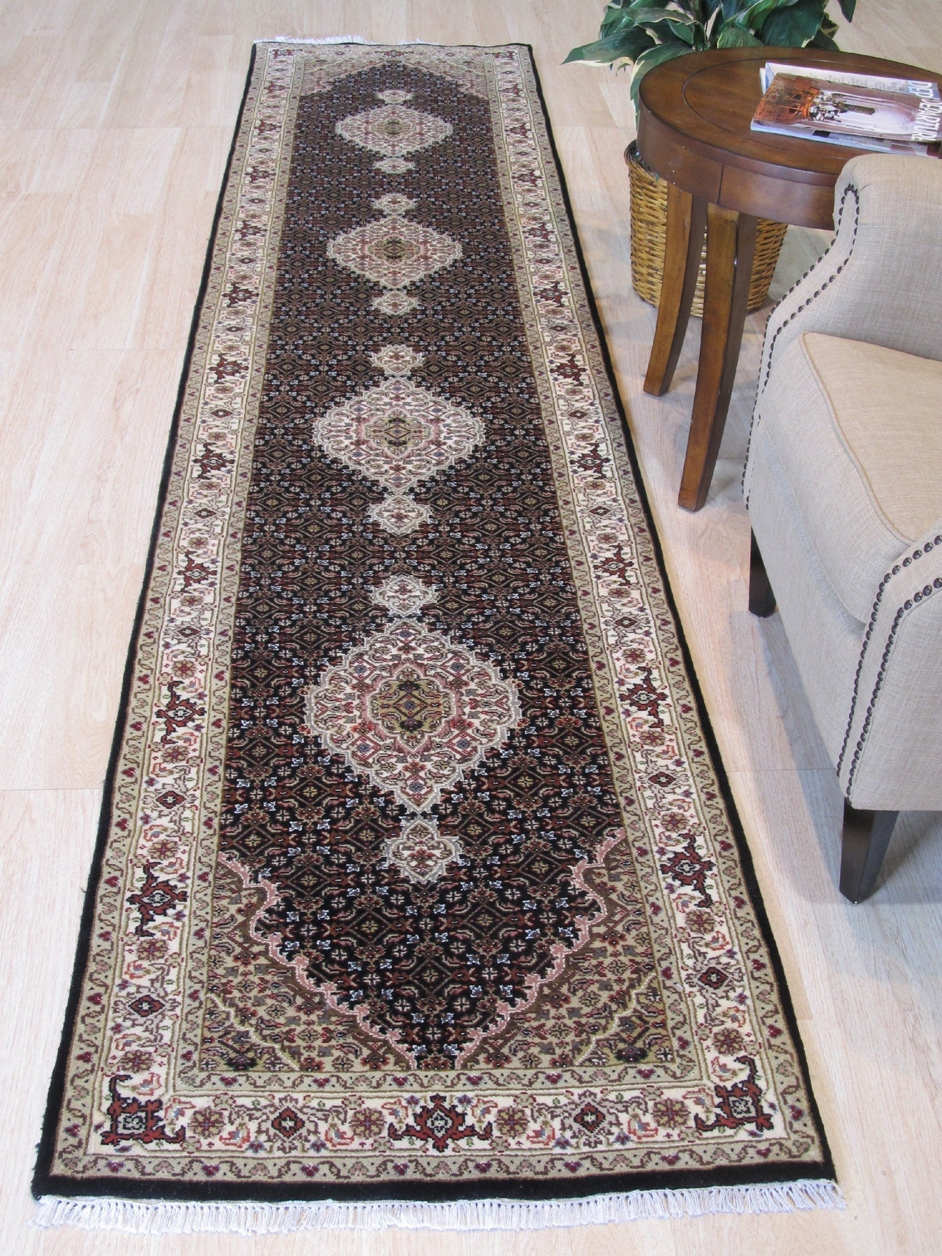 Ophelia Amara Brown Hand Knotted Wool Rug, displaying intricate patterns in shades of brown and cream, lays flat on a light wooden floor, its hand-knotted texture subtly visible.