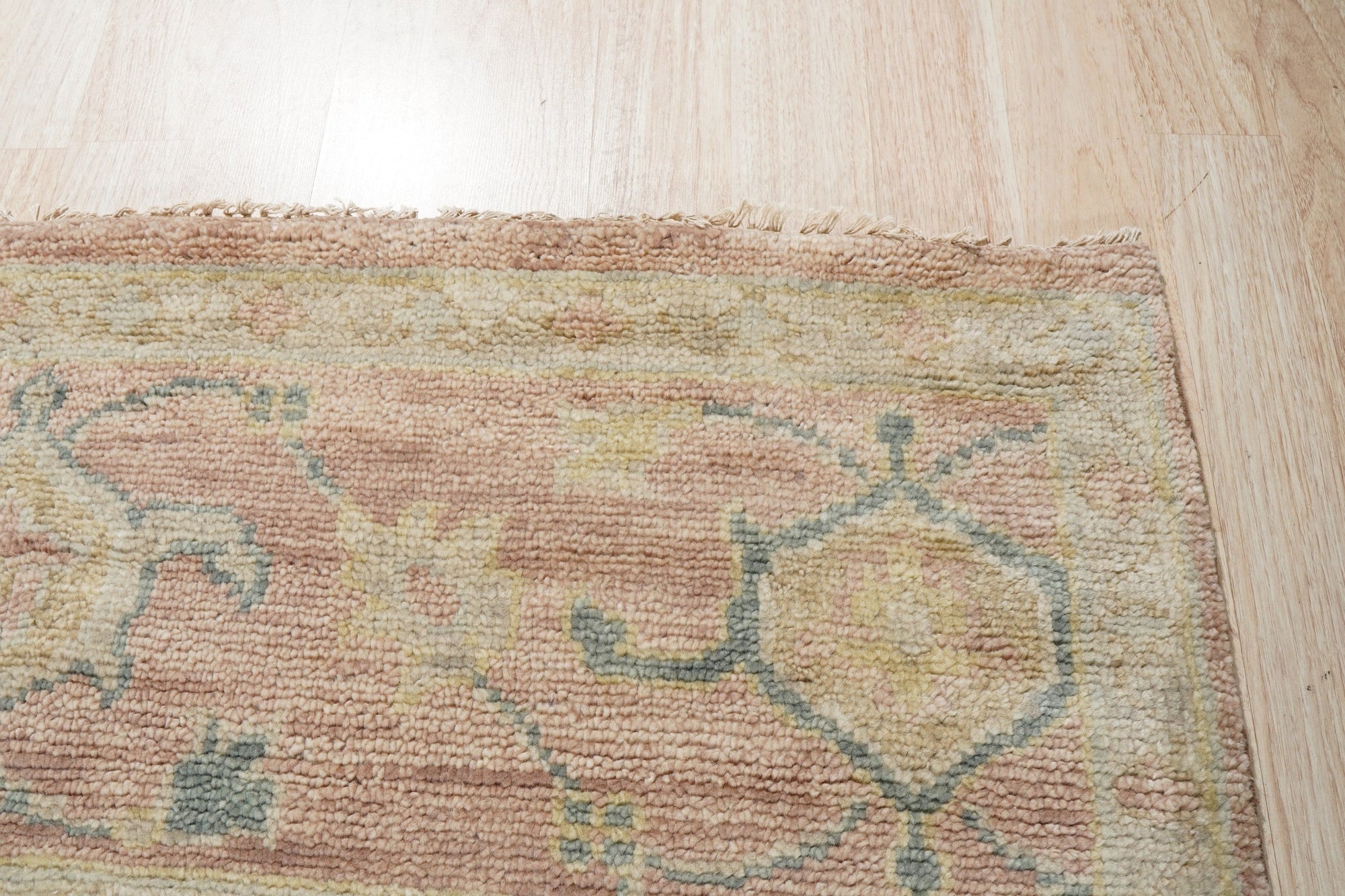 Timeless Oushak Pink Hand Knotted Wool Rug, displaying a delicate floral pattern in soft pink hues, graces a bright living space with warm wooden floors, hinting at classic elegance and handcrafted comfort.