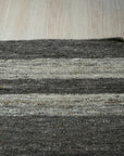 Cassandra Clara Brown Flatweave Wool Rug, a warm brown wool rug with a subtle geometric pattern, complementing a bright, minimalist living room with light wood flooring and a sleek black accent chair.