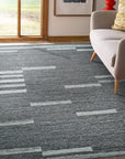 Cassandra Clara Brown Flatweave Wool Rug graces a warmly lit room; its flatweave construction showcasing a sophisticated, muted brown palette complementing natural wood furniture and creating a serene, inviting atmosphere.