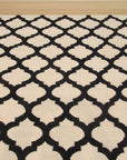 Maya Moroccan Black Flatweave Wool Rug, seen in a living room setting, its geometric black pattern contrasting against light wood floors and a neutral sofa.