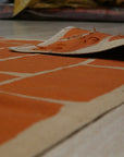 Kira Amara Orange Flatweave Wool Rug, showcasing a vibrant orange and cream geometric pattern in a modern living room setting. Light wood floors and a neutral sofa complement the rug's warm tones.