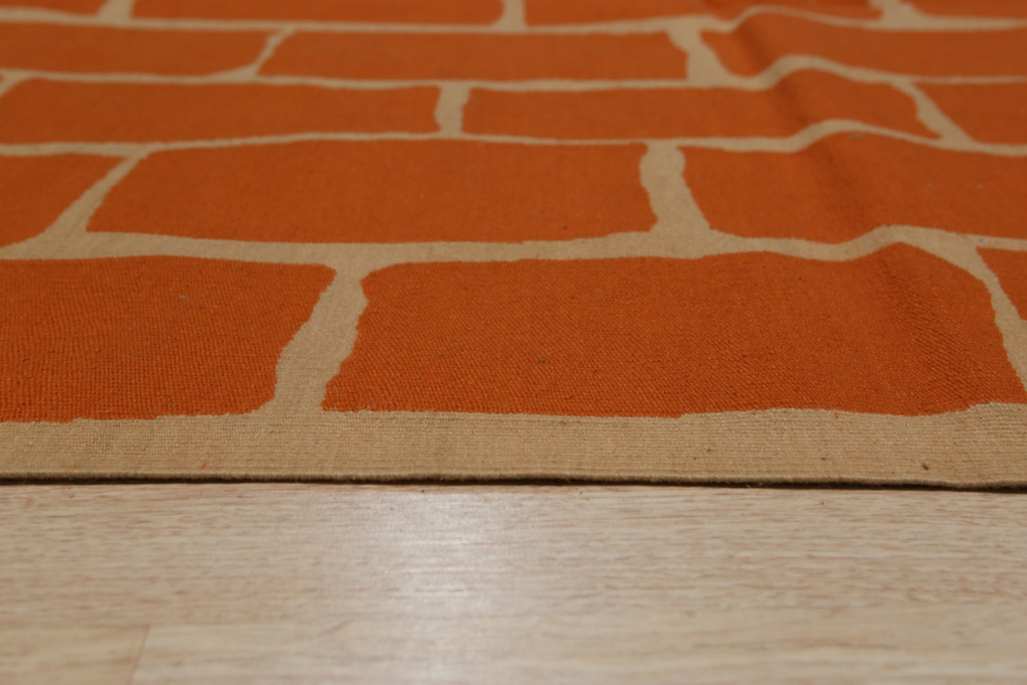 Kira Amara Orange Flatweave Wool Rug, a vibrant geometric pattern in shades of orange and cream, lays flat on a light wood floor, its textured wool visible in the bright, natural light of a modern living space.