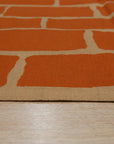 Kira Amara Orange Flatweave Wool Rug, a vibrant geometric pattern in shades of orange and cream, lays flat on a light wood floor, its textured wool visible in the bright, natural light of a modern living space.