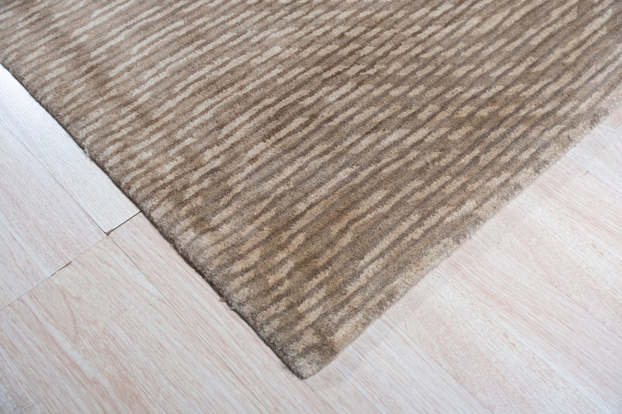 Sloane Freya Brown Hand Tufted Wool Rug, a warm brown and cream rug with a subtle geometric pattern, lies elegantly on a light hardwood floor, partially beneath a dark wood coffee table, showcasing its plush texture and grounding presence in a living space.