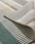 Zinnia Striped Green Handmade Wool Rug displayed in a bright, airy living space. Natural light highlights the rug's vibrant green and neutral stripes, showcasing its textured wool construction and handmade quality. The rug anchors a contemporary seating area, lending a fresh, organic feel to the room.
