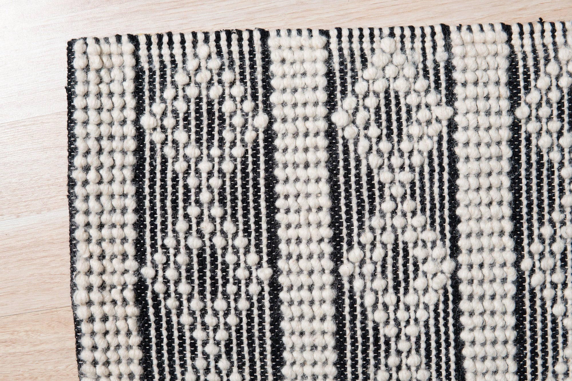 Selene Striped Black Handmade Wool Rug, a textural focal point in a bright, modern living room. Thick black and off-white stripes run horizontally across the wool rug, grounding a space with light wood furniture and a neutral palette.