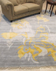 Sloane Giselle Gray Hand Knotted Wool & Viscose Rug, a luxurious blend of wool and viscose, rests on a dark wood floor, its subtle gray tones and intricate hand-knotted design creating a sophisticated and calming atmosphere within a softly lit room.