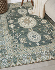 Rhea Traditional Green Hand Knotted Wool Rug, displaying intricate floral patterns in shades of green, cream, and hints of red, gracing a light hardwood floor under soft, diffused light.