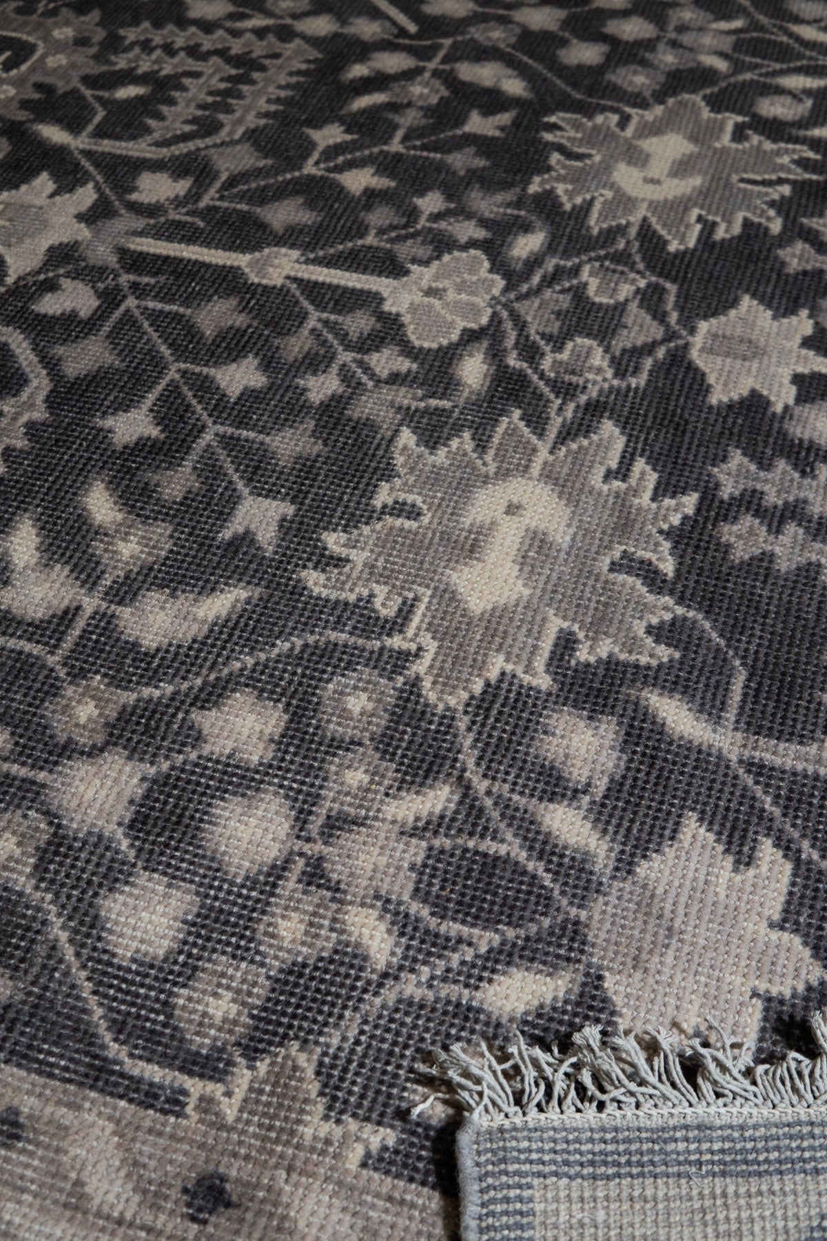 Cozy Oushak Charcoal Hand Knotted Wool Rug, a charcoal wool rug with a traditional Oushak design, lies partially rolled out on a light-colored floor, showcasing its texture and intricate patterns.