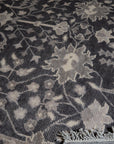 Cozy Oushak Charcoal Hand Knotted Wool Rug, a charcoal wool rug with a traditional Oushak design, lies partially rolled out on a light-colored floor, showcasing its texture and intricate patterns.