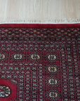 Rustic Nina Red Nan Rug, a vibrant geometric pattern in warm reds and creams, lies flat on a polished concrete floor. Tassels fringe one end, hinting at a hand-woven construction. Subtle variations in texture and color add rustic charm.