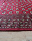Rustic Nina Red Nan Rug, its textured weave in warm reds and earthy tones, partially visible in a warmly lit room, suggesting cozy comfort and rustic charm.