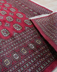 Rustic Nina Red Nan Rug sprawls across a warmly lit wooden floor, its rich red tones and intricate, distressed pattern evoking a sense of aged elegance. Light catches the textured surface of the Nan-style weave.