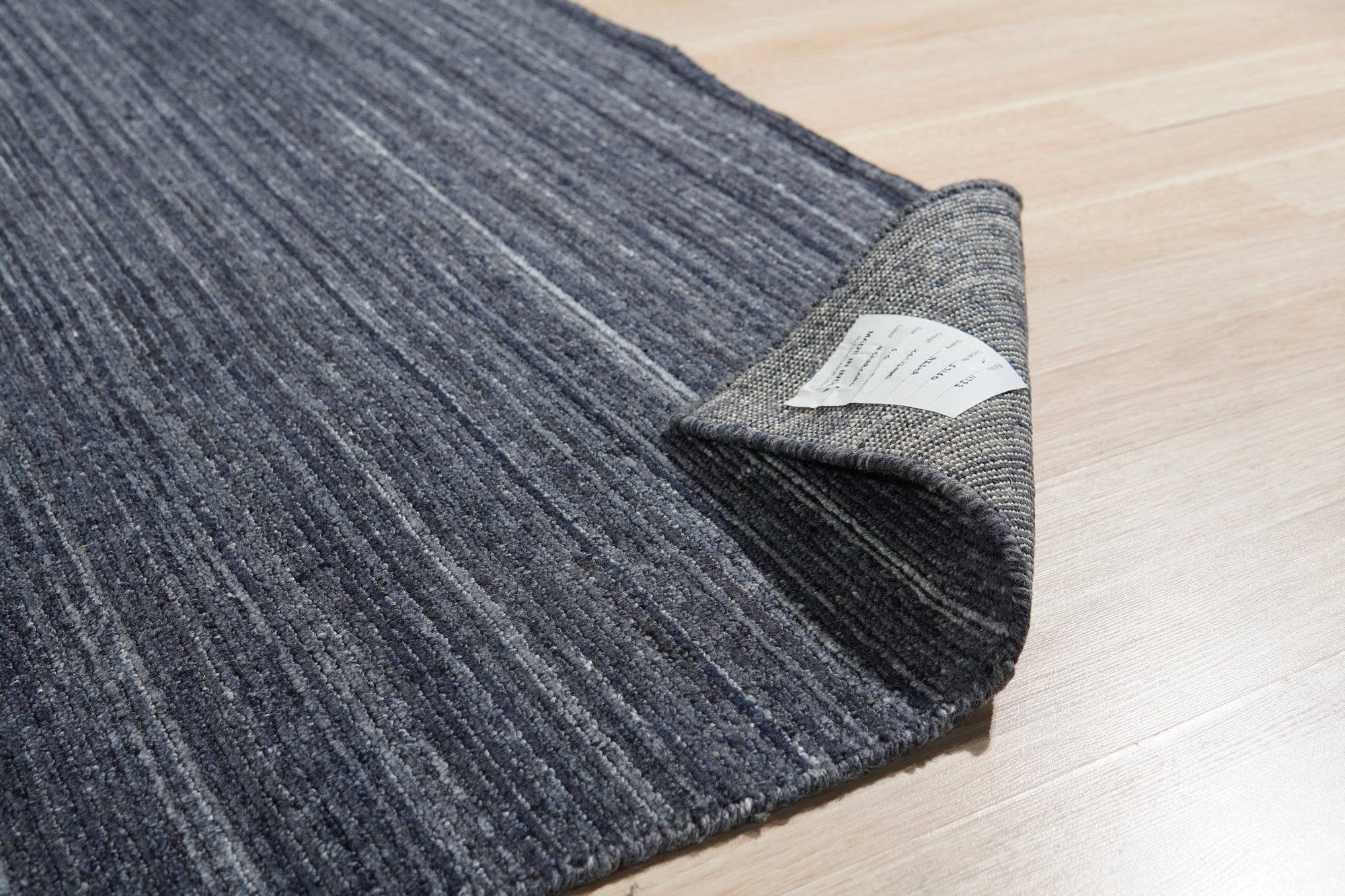Delicate Striped Charcoal Handmade Wool Rug, displays narrow stripes of varying charcoal and grey tones woven in wool, adding a subtle textural element to a warmly lit room with hardwood floors.