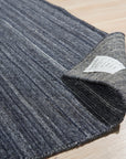 Delicate Striped Charcoal Handmade Wool Rug, displays narrow stripes of varying charcoal and grey tones woven in wool, adding a subtle textural element to a warmly lit room with hardwood floors.