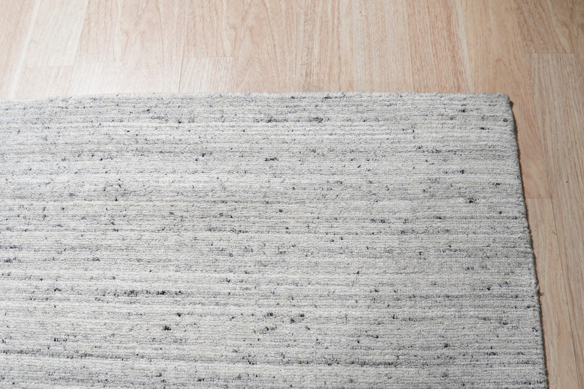 Delicate Striped Silver Handmade Wool Rug, showcasing subtle silver stripes across a textured wool weave, sits elegantly on a light hardwood floor. Natural light illuminates the rug's soft sheen.