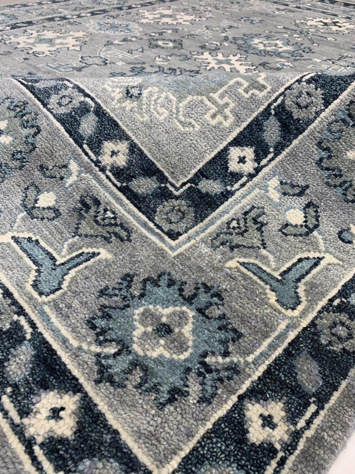 Maya Oushak Silver Hand Knotted Wool Rug, showcasing a silvery-grey palette and intricate floral patterns woven in hand-knotted wool, lies gracefully on the floor in a warmly lit interior space.
