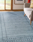 Jasmine Alluring Gray Handmade Wool Rug displayed in a softly lit living room. Gray hues blend across its wool texture, contrasting subtly with a light wooden floor. Corner of a mid-century modern chair adds scale.