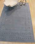Jasmine Alluring Light Blue Handmade Wool Rug graces a bright, stylish living room. Soft light washes over the rug’s intricate pattern and gentle light blue hue, highlighting its handcrafted wool texture beneath a minimalist coffee table and modern furniture.