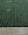 Laine Amara Green Handmade Wool Rug, seen in a bright, inviting living room. The rug's textured wool pile adds warmth to the light wood flooring, complementing the neutral furniture and potted plant.