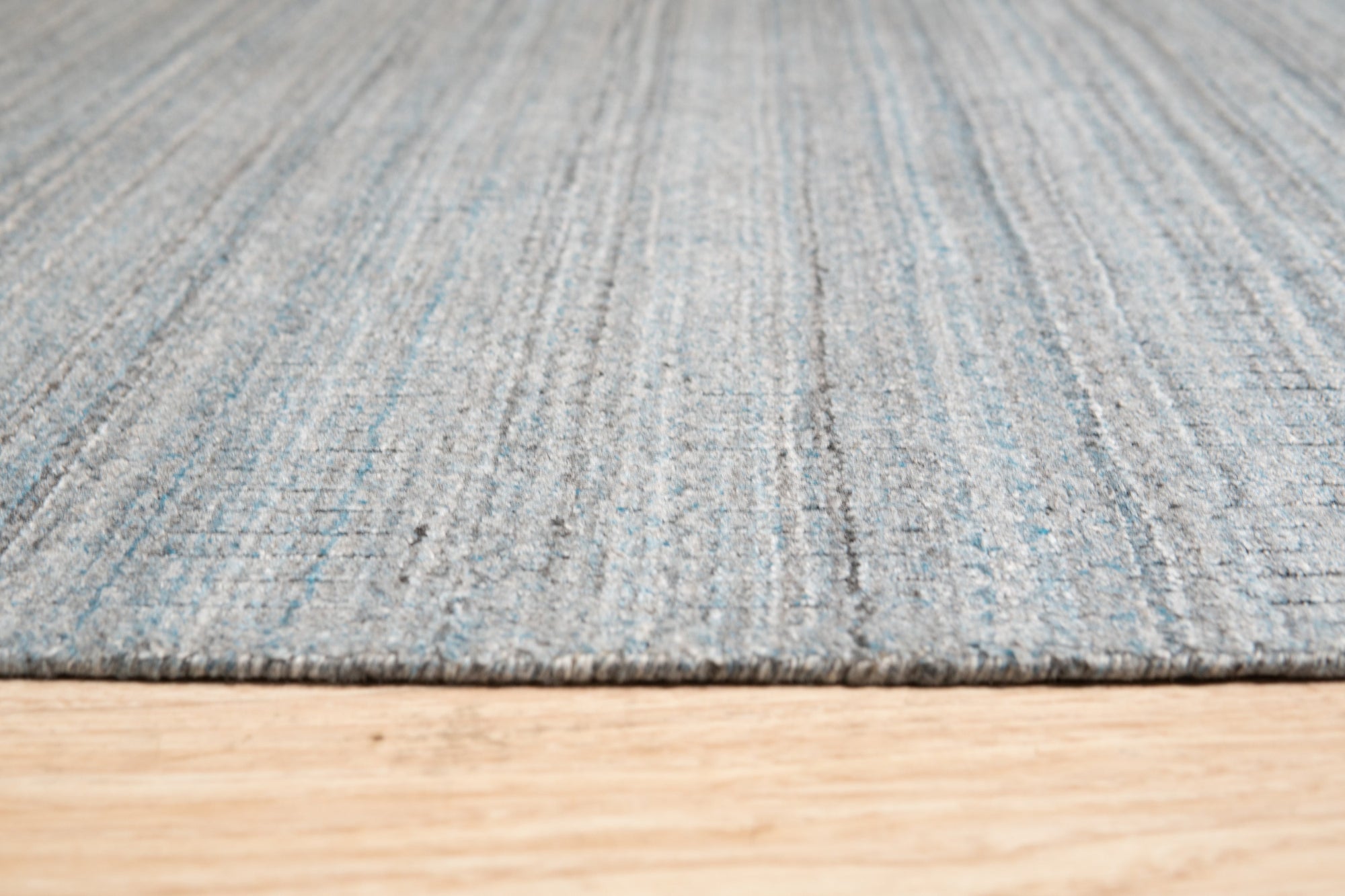 Para Elena Blue Handmade Wool Rug, featuring intricate geometric patterns in varying shades of blue, grey, and cream, laid on a light hardwood floor bathed in soft, natural light; corner view showcasing handcrafted wool texture and artisanal detail.