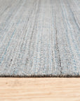 Para Elena Blue Handmade Wool Rug, featuring intricate geometric patterns in varying shades of blue, grey, and cream, laid on a light hardwood floor bathed in soft, natural light; corner view showcasing handcrafted wool texture and artisanal detail.