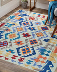 Nina Cassandra Multicolor Flatweave Wool Rug, vibrant geometric patterns in blues, oranges, and yellows dance across this wool rug, seen in a modern living room setting with a mid-century style chair.