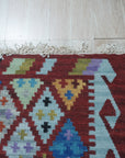 Amara Kilim Multicolor Flatweave Wool Rug, vibrant hues dance across a flatwoven wool surface, creating a dynamic geometric pattern in a well-lit interior space.
