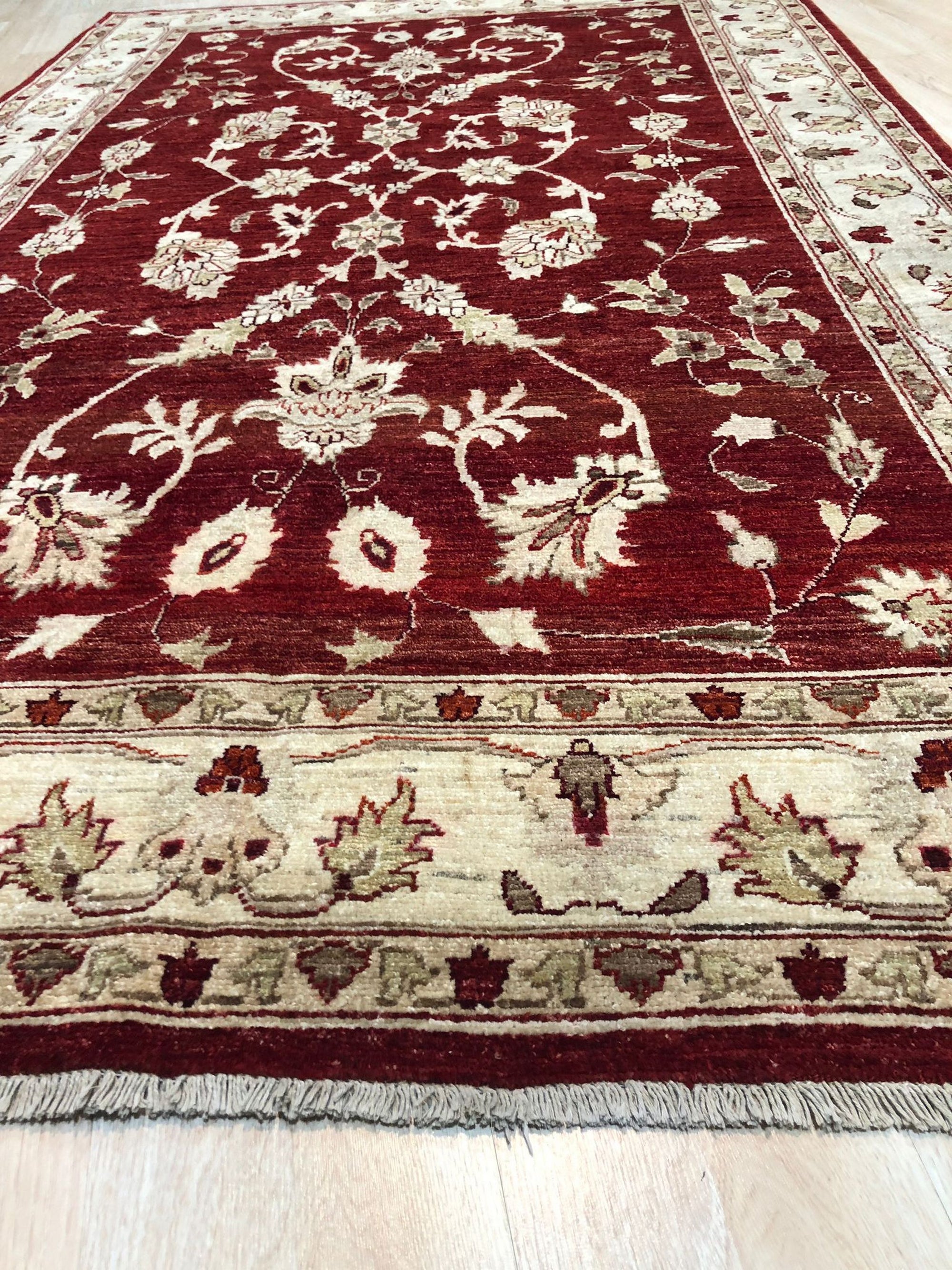 Rustic Agra Red Hand Knotted Wool Rug, showcasing its intricate floral patterns in warm reds and creams, lies gracefully on a light hardwood floor, adding a touch of timeless elegance to the living space.