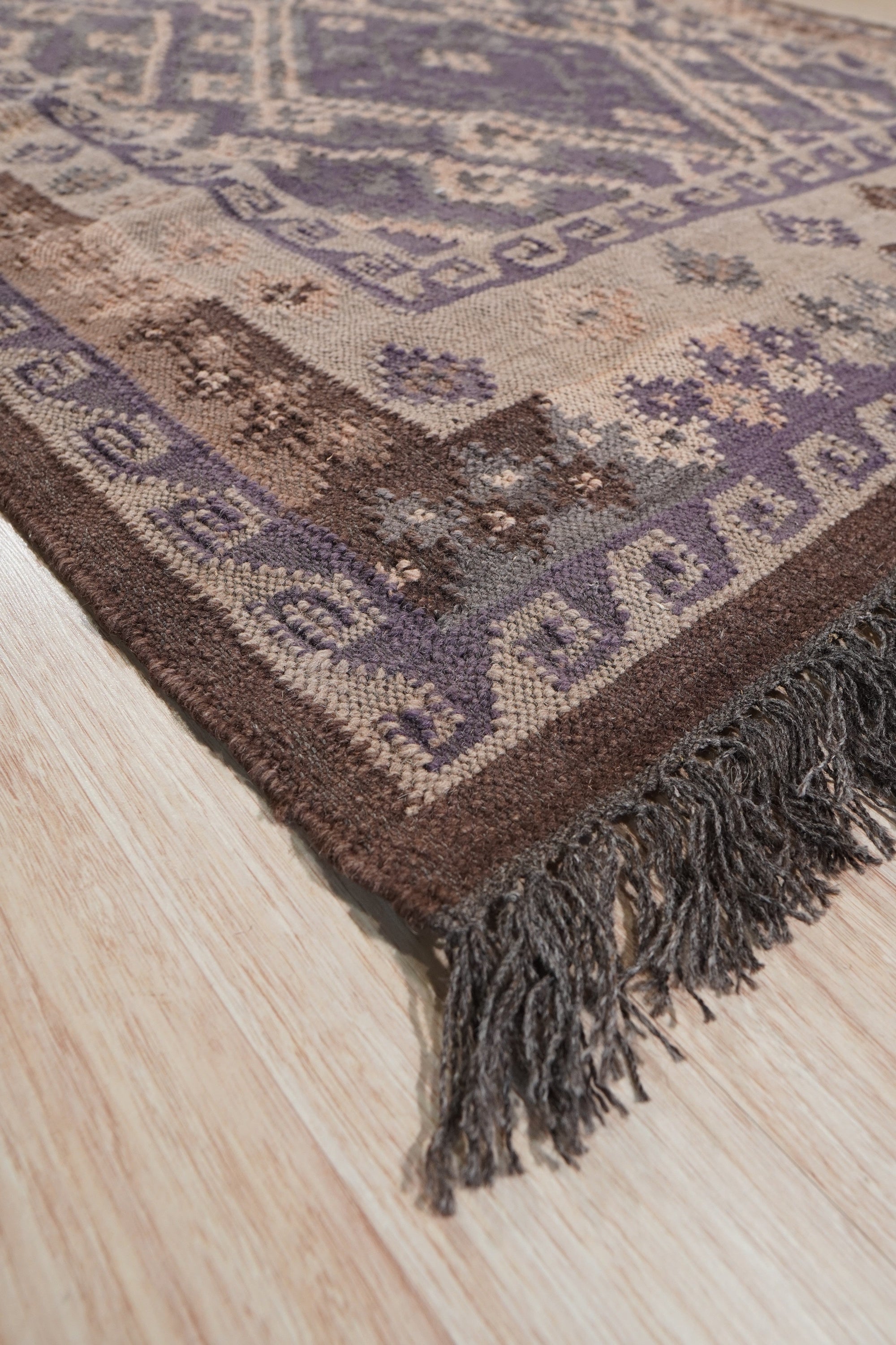Brenda Jasmine Brown Flatweave Wool Rug, displaying its intricate geometric pattern in warm browns and creams, lies flat on a light wood floor, showcasing its soft, textured wool construction.