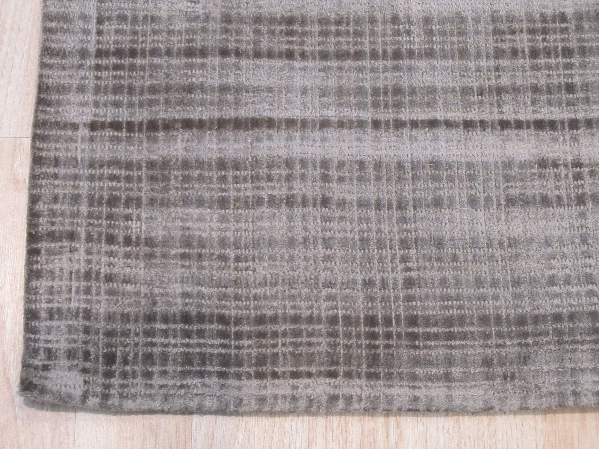 Sloane Maya Silver Handmade Rug gracing a living room, its subtle silver hues and textural weave complement a modern grey sofa and sleek wooden coffee table. Warm light illuminates the rug's intricate pattern, creating a cozy and inviting atmosphere.