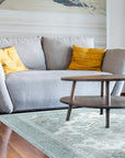 Selene Oriental Green Handmade Wool Rug graces a warmly lit living room. The hand-knotted wool rug, a vibrant blend of greens and earthy tones, anchors a space with a mid-century modern aesthetic. Sunlight highlights the intricate oriental pattern and plush texture under a stylish coffee table.