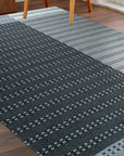 Clara Xena Charcoal Flatweave Wool & Cotton Rug, a charcoal and ivory patterned rug, anchors a modern living room with a light wood coffee table and cozy throw pillows.