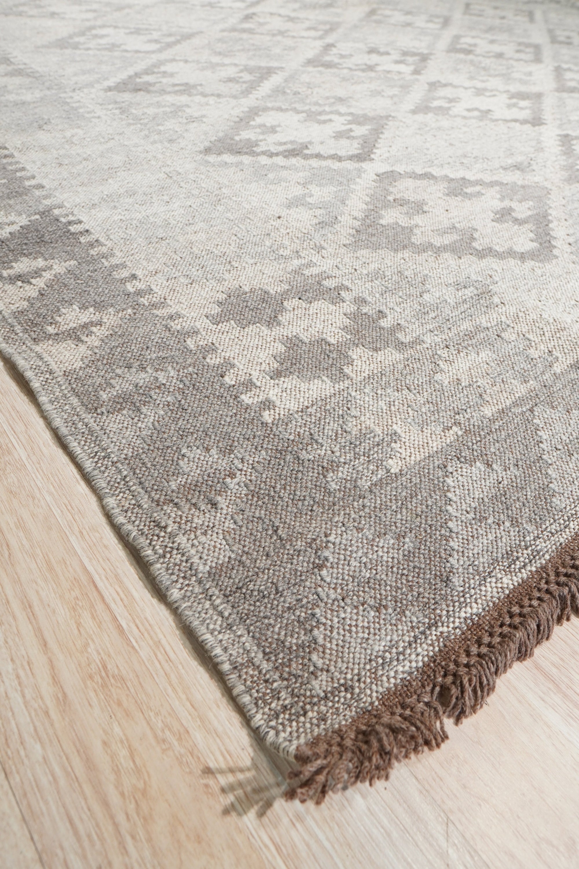 Brenda Para Gray Flatweave Wool Rug, displaying subtle geometric patterns in shades of gray, lies flat on a light wood floor in a brightly lit room, showcasing its texture and minimalist design.