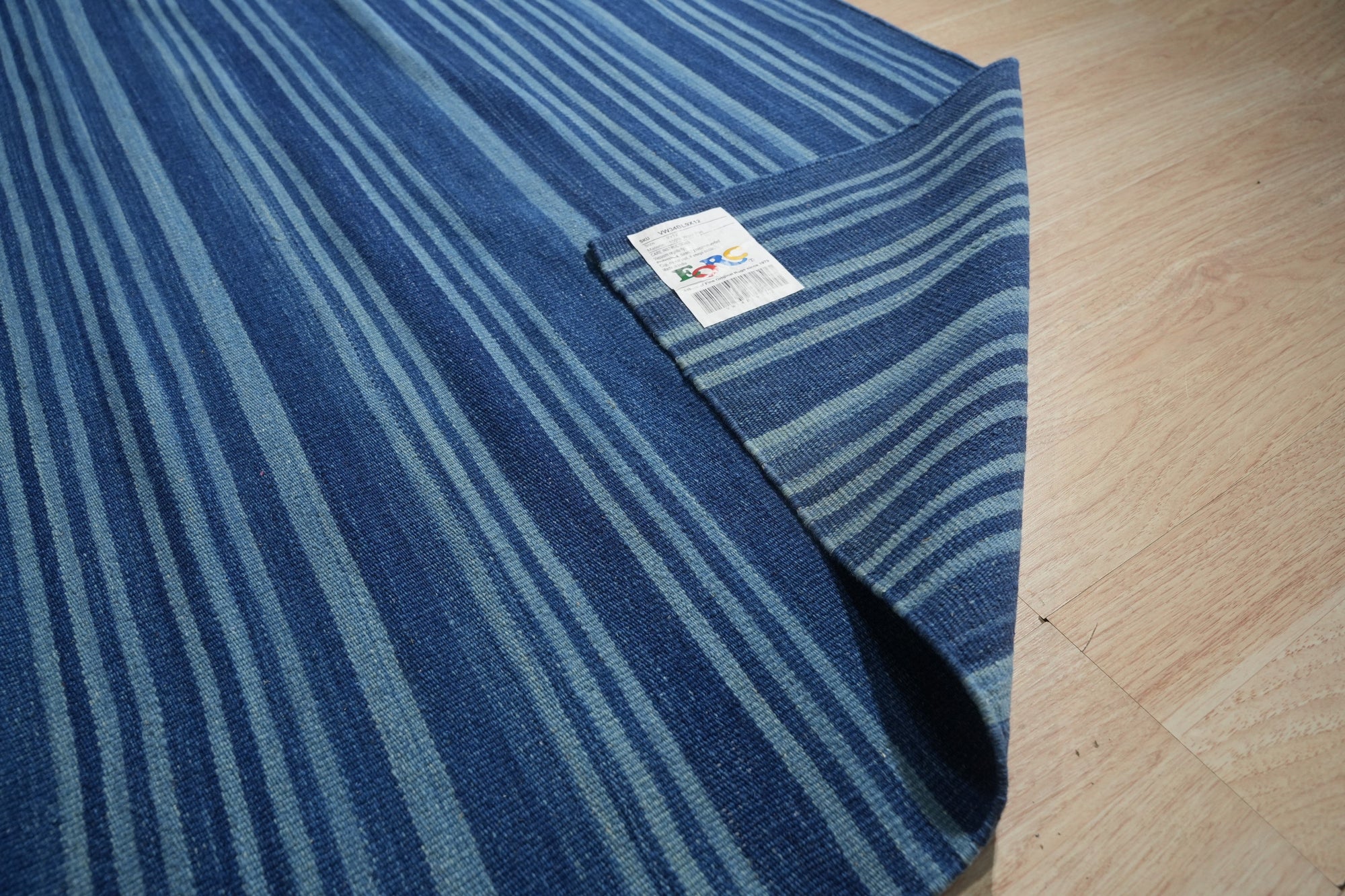 Para Penelope Blue Flatweave Wool Rug, showcasing a geometric pattern with varying shades of blue and cream, lies on a light-colored wooden floor, partially obscured by a modern gray sofa with wooden legs and a woven basket in the background, hinting at a comfortable, contemporary living space.