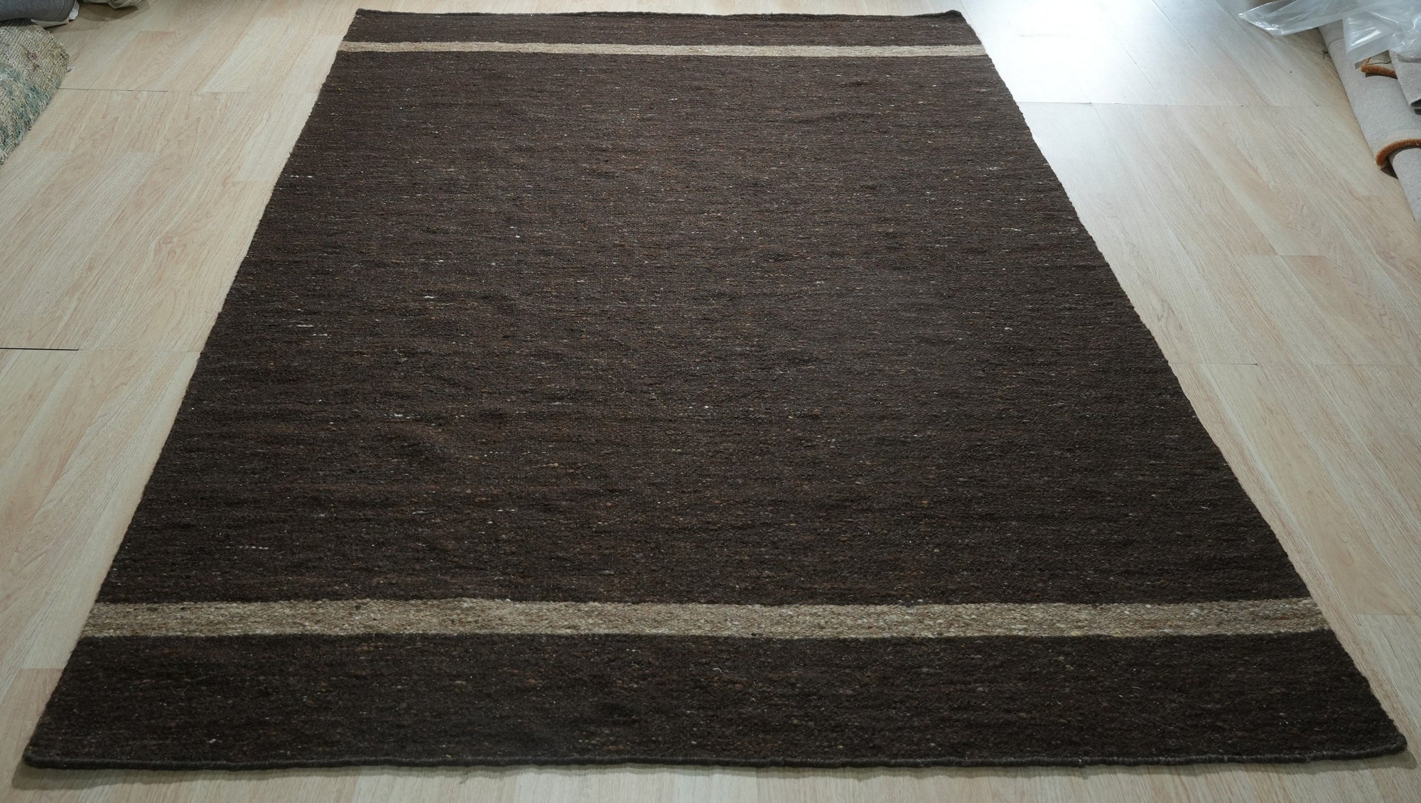 Violet Artistic Brown Flatweave Wool Rug sprawls in a sun-drenched living room, its earthy brown hues and artistic design complementing a modern sofa and textured throw pillows. Flatweave construction offers a low profile against a light wood floor, creating a warm and inviting space.
