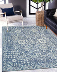 Ava Agra Blue Hand Knotted Wool & Bamboo Silk Rug, a blend of calming blues and shimmering silk highlights, lies subtly beneath a mid-century modern styled living room with light gray walls, dark brown hard wood flooring, and an accent chair in the background.