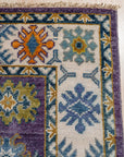 Cassandra Kazak Purple Hand Knotted Wool Rug, a rich purple and wool rug featuring a traditional Kazak design, fills the frame with its intricate geometric patterns.