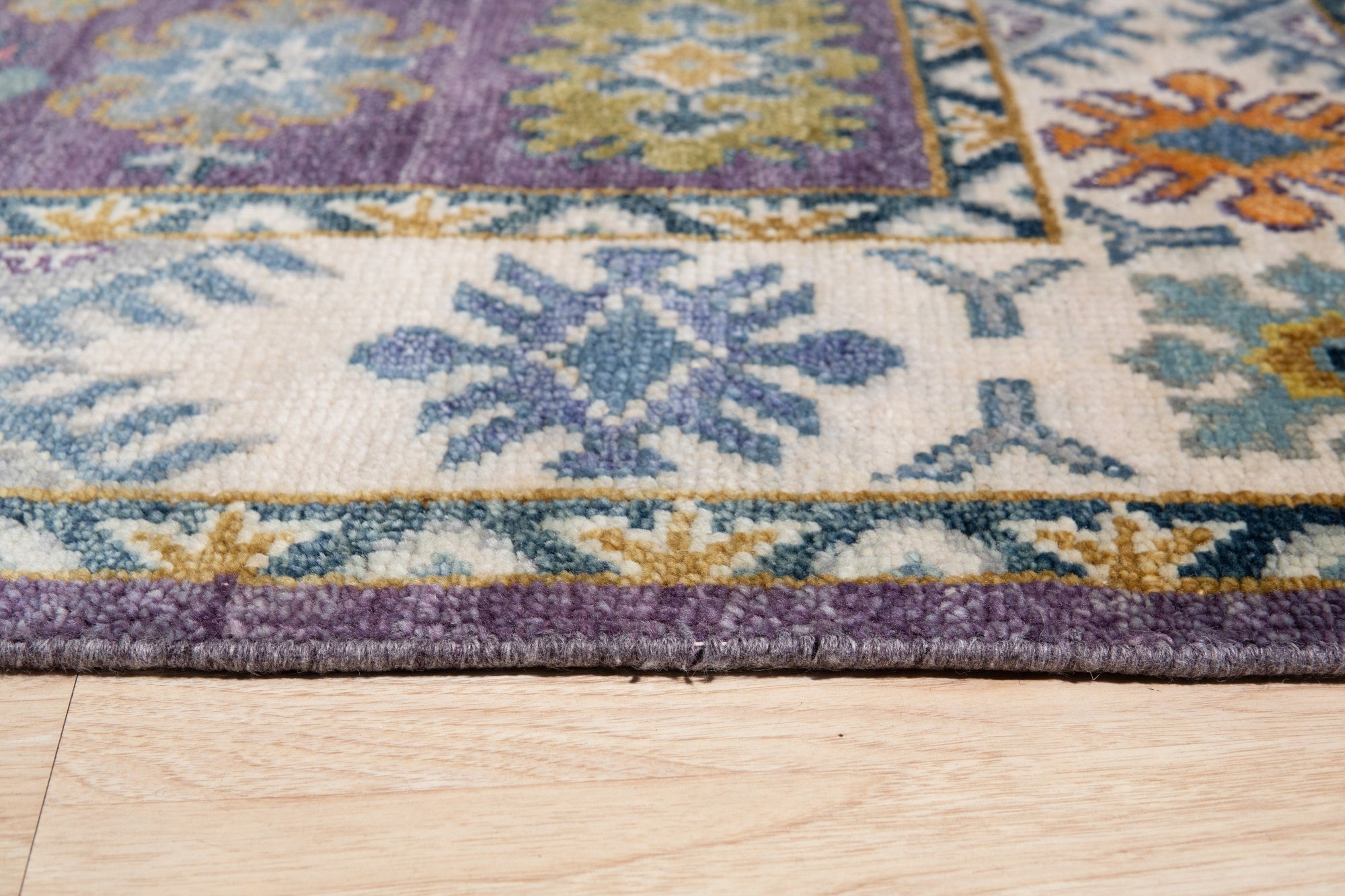 Cassandra Kazak Purple Hand Knotted Wool Rug, showcasing intricate geometric patterns in shades of purple, red, and cream, lies on a light wood floor with natural light illuminating its plush wool texture.