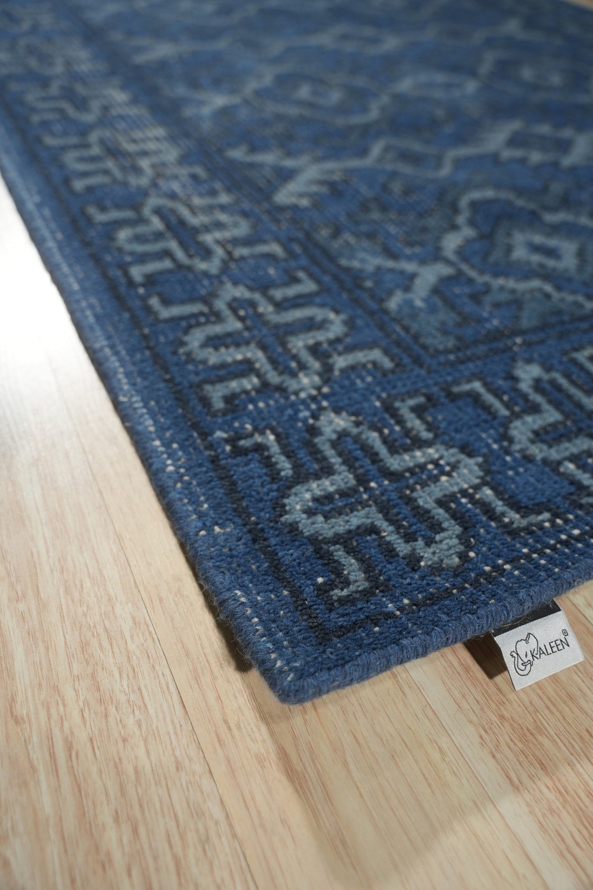 Mira Traditional Blue Hand Knotted Wool Rug, displays a complex, geometric pattern in varying shades of blue, ivory, and hints of red, lending a sophisticated touch to the warmly lit living room setting.